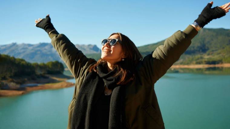 A woman standing in nature holds her arms up and smiles