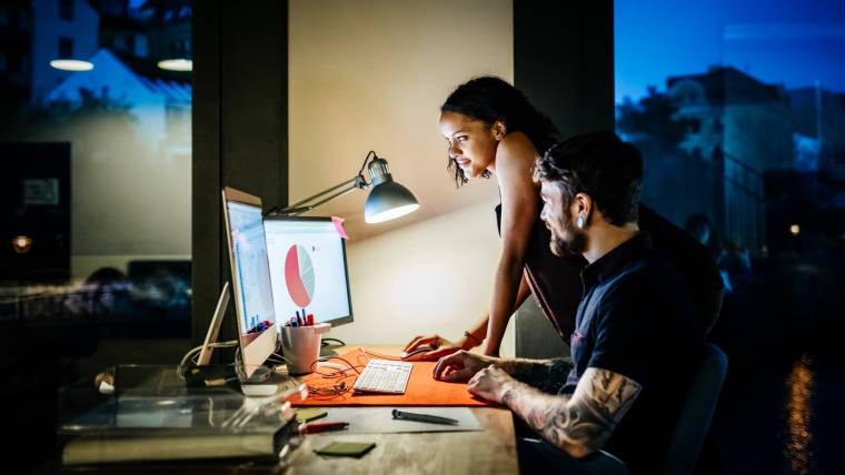 Two workers smile at computer screens with graphs on them