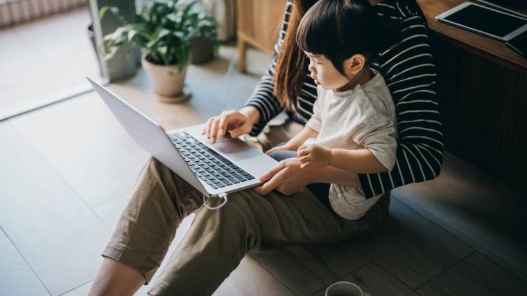 a working mom holds her toddler while looking at her laptop