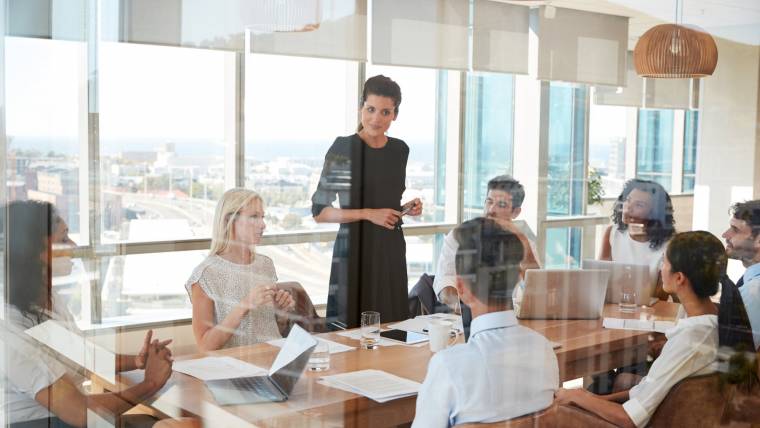 A business leader stands over a conference table and listens to a colleague