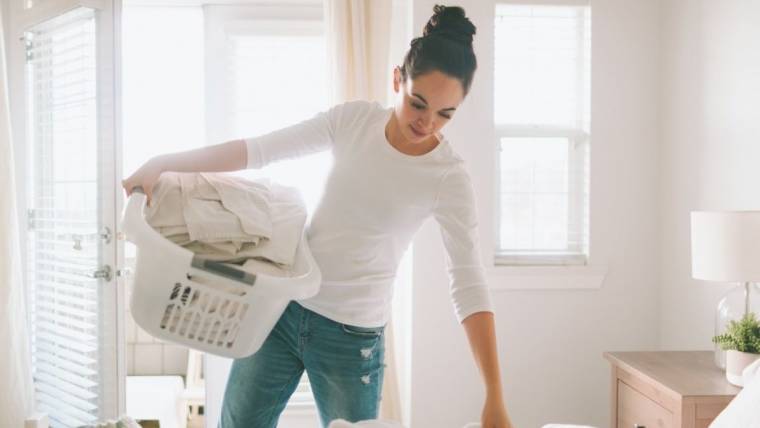 A mom does holds a basket of laundry against her hip while making a bed