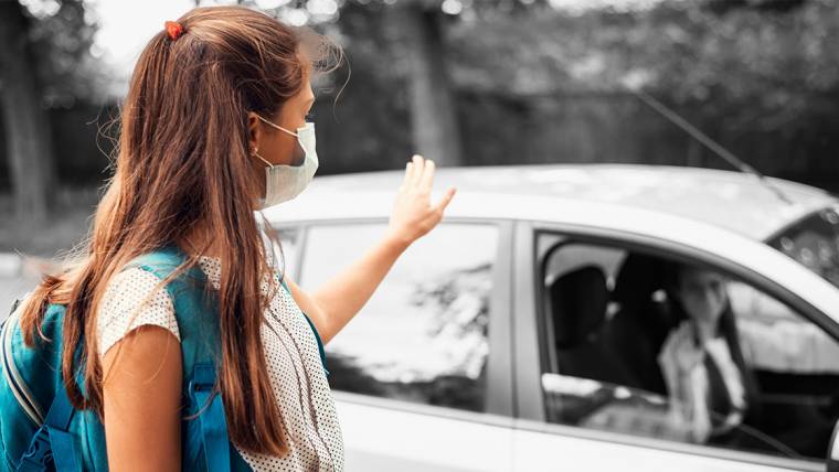 masked child wearing a backpack and waving goodbye to parent after being dropped off at school