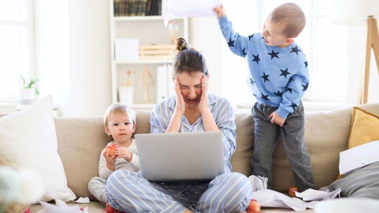 woman in pajamas looking stressed while holding a laptop in her lap and siting with two small children