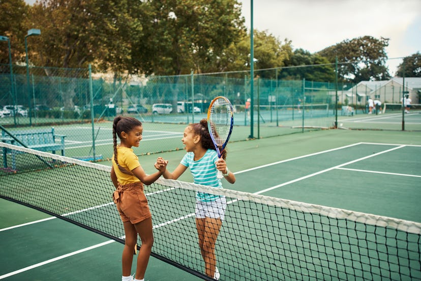 Two young girls shake hands and smile over the net on a tennis court, in a story about sports kids c...