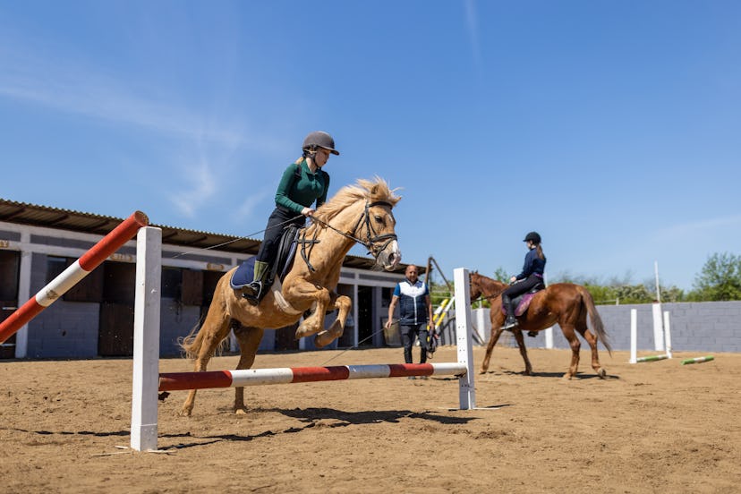 Two girls riding horses together outdoors as one goes over a jump, in a story about sports kids can ...